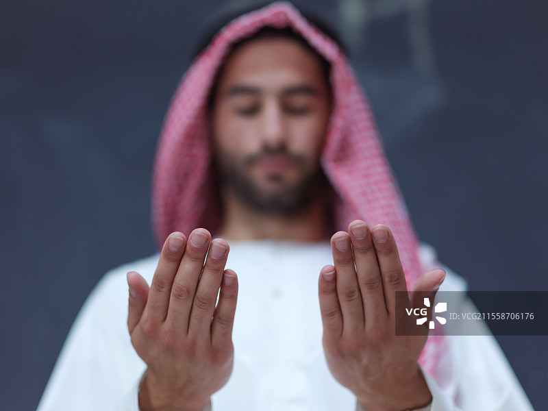 年轻的阿拉伯男子穿着传统服装，双手合十在黑色黑板前祈祷，展现现代伊斯兰时尚和斋月的概念。图片素材