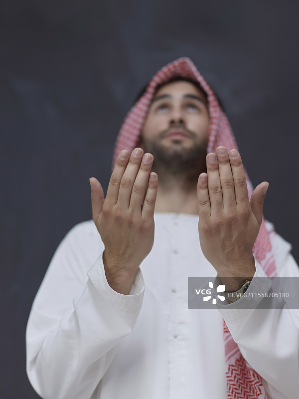 身穿传统服装的年轻阿拉伯男子在黑色黑板前以祈祷姿势向上帝祈祷，体现现代伊斯兰时尚和斋月的概念。图片素材