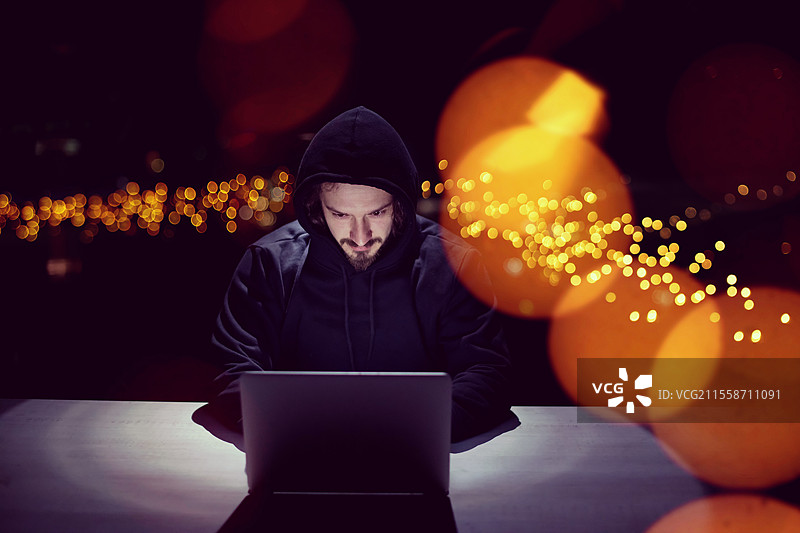 年轻有才华的黑客在黑暗的办公室里使用笔记本电脑，背景是夜晚的大城市灯光图片素材