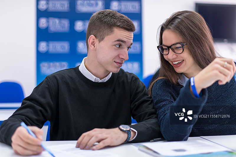 两名年轻学生（男生和女生）与其他同学在教室里一起进行项目，写笔记并讨论。图片素材
