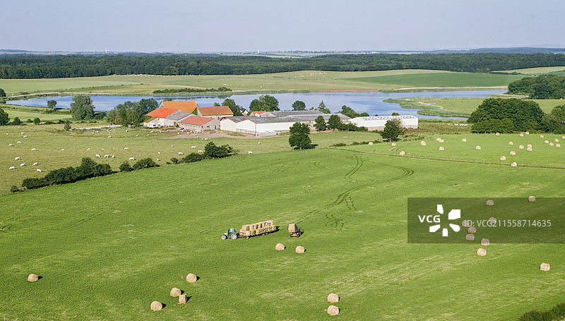 干草收割的鸟瞰图。将干草卷装载到拖车上。位于池塘边缘的孤立农场。法国洛林。图片素材