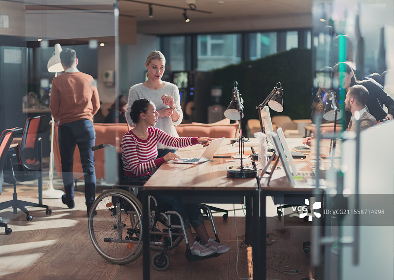 坐在轮椅上的残疾女商人在创意办公室工作。现代共享办公空间中的商业团队。背景中同事们在深夜工作。包容性和残疾概念。坐在轮椅上的残疾女商人在现代共享办公空间中。背景中的同事们。残疾和障碍概念。选择性聚焦。图片素材