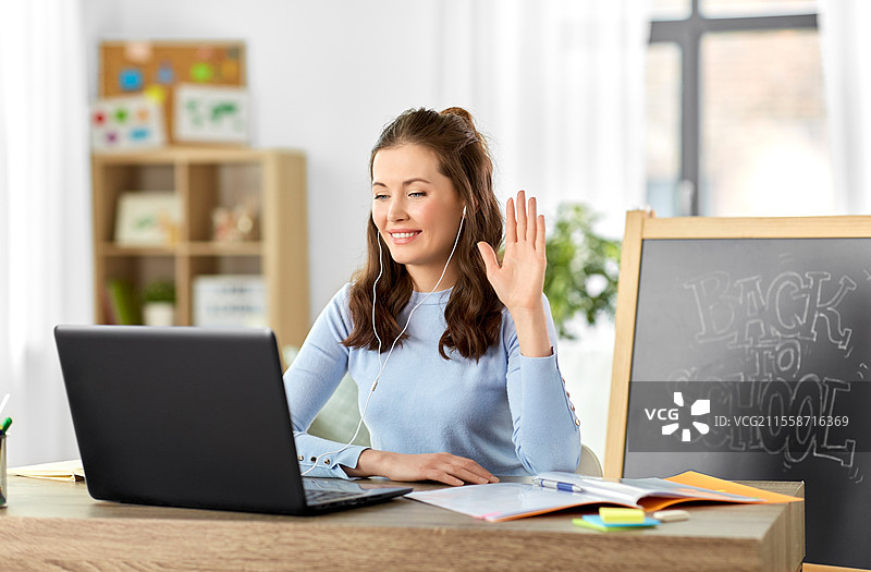 远程教育、远程工作和人们的概念 - 开心微笑的女教师在家办公室使用笔记本电脑和耳机进行在线课程或视频通话，并向镜头挥手。女教师在家中使用笔记本电脑进行在线课程。图片素材