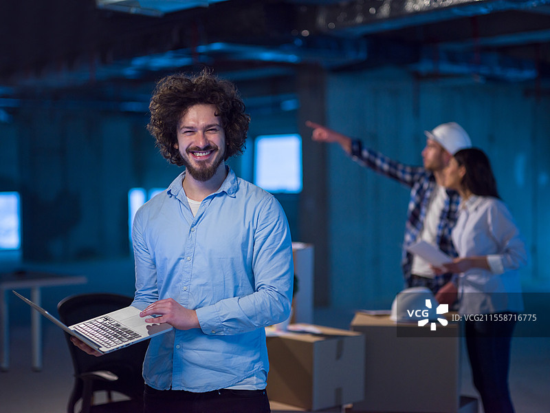 年轻男性建筑师在施工现场查看文件和商业流程，背景中有同事们在新创办的办公室里使用笔记本电脑。图片素材