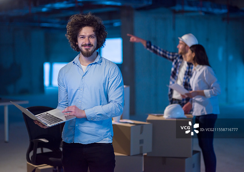 年轻男性建筑师在施工现场查看文件和商业流程，背景中有同事们在新创办的办公室里使用笔记本电脑。图片素材