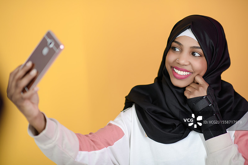 一位面带美丽微笑的非洲穆斯林女性在黄色背景下用手机自拍图片素材
