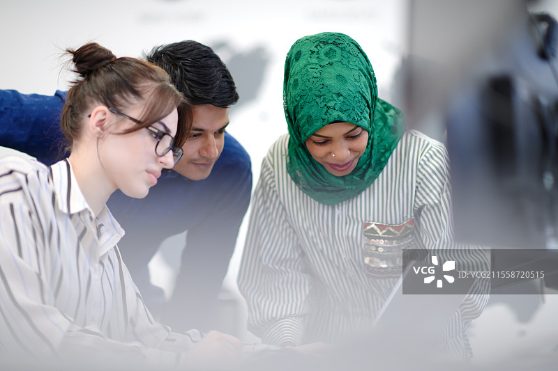 多元文化的初创企业团队，阿拉伯女性戴着头巾，在现代开放式办公室内开会，进行头脑风暴，使用笔记本电脑和台式电脑工作。图片素材