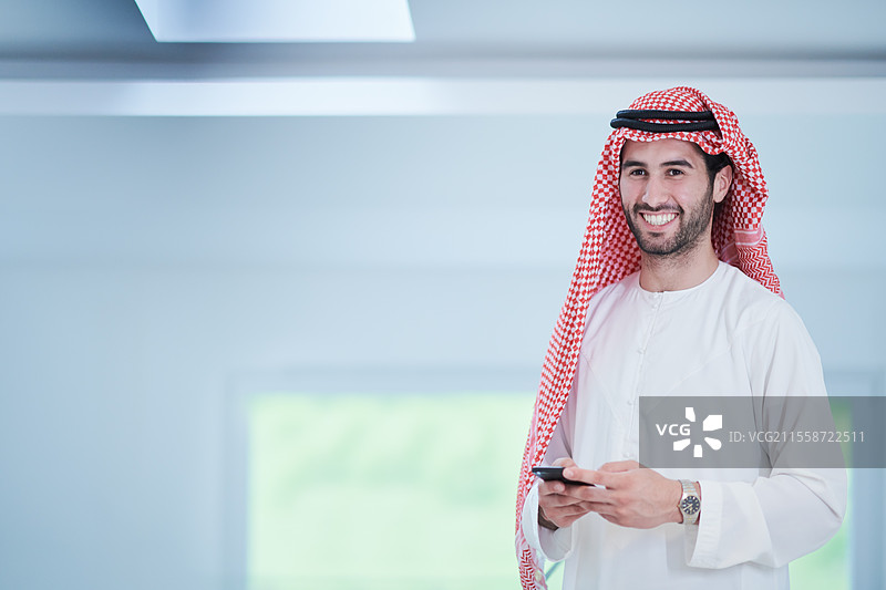 年轻的现代穆斯林阿拉伯商人穿着传统服装，在家中使用智能手机图片素材