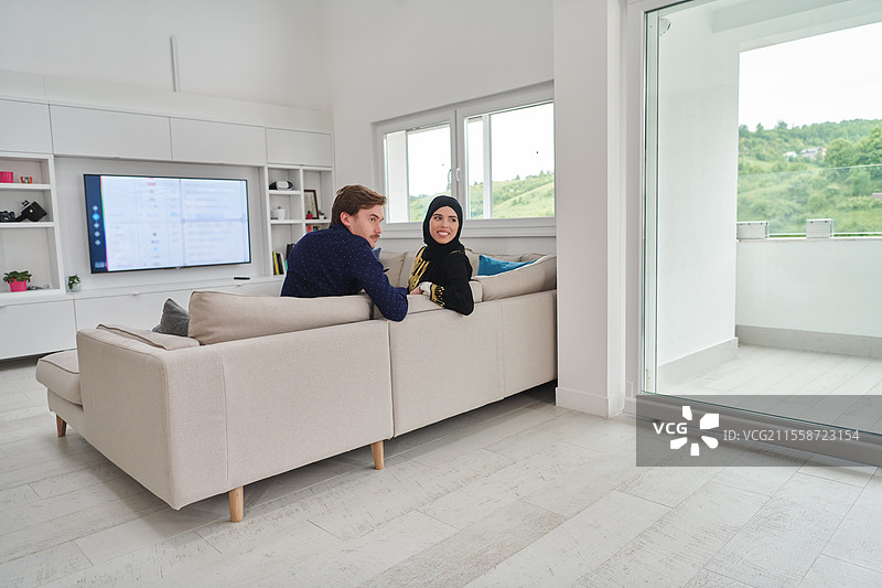 年轻的穆斯林夫妇在斋月期间享受在家时光。快乐的阿拉伯家庭在看电视。图片素材