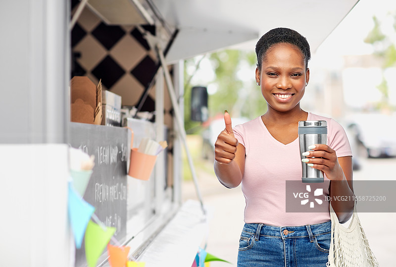 可持续发展与人文概念 - 年轻的非裔美国女性手持保温杯和装在绳袋里的食物，背景是食品车，竖起大拇指。图片素材