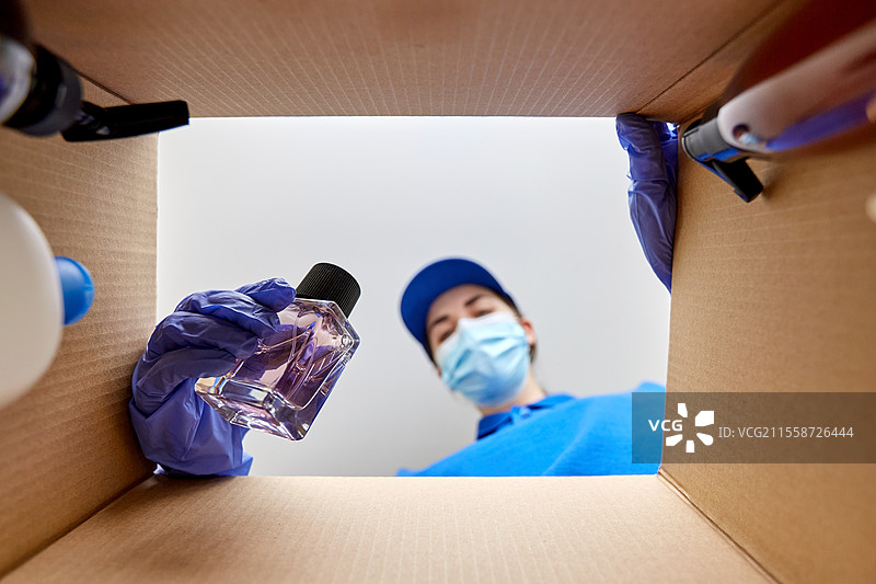 家庭配送、运输和疫情概念 - 戴着防护口罩和手套的女性正在打包装有香水和美容产品的包裹盒。戴着口罩的女性正在打包装有化妆品的包裹盒。图片素材