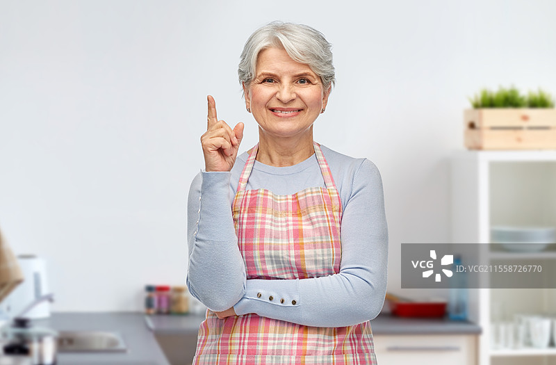 烹饪、烹饪艺术和老年人概念 - 微笑的老年女性穿着围裙，手指向上，背景是家里的厨房。微笑的老年女性在厨房里手指向上。图片素材