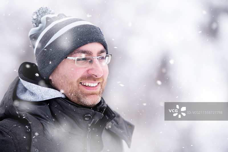 冬季的年轻男子肖像，穿着温暖的衣服，享受着雪花飘落的雪天，身处冬季森林中。图片素材