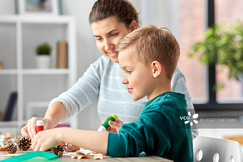家庭、创造力和手工艺概念 - 母亲和小儿子在家用胶水和纸制作干燥秋叶、松果和栗子的图片。母亲和儿子正在制作秋叶的图片。图片素材