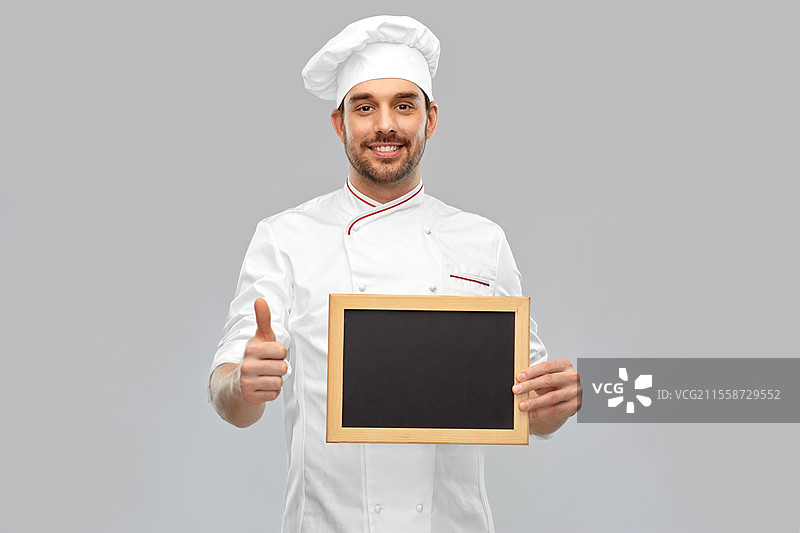 烹饪、烹饪艺术和人们的概念 - 开心微笑的男厨师戴着厨师帽，在灰色背景前展示黑板并竖起大拇指。开心的男厨师展示黑板并竖起大拇指。图片素材