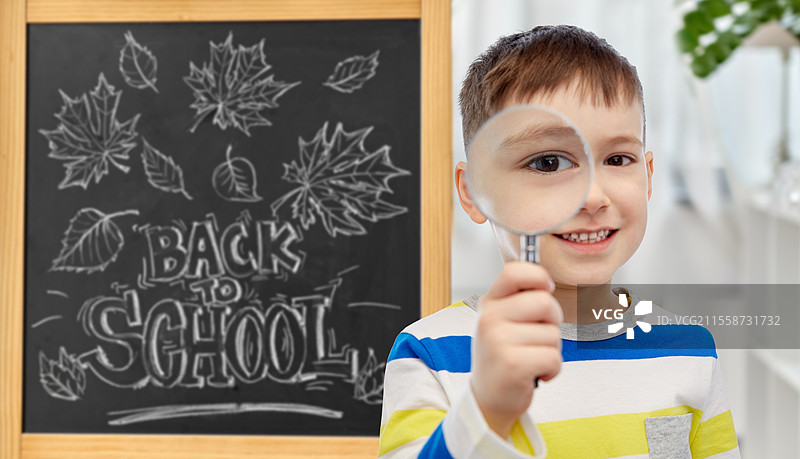 教育、学习和人们的概念 - 快乐的小学生男孩透过放大镜看黑板，背景是“开学”字样。学生男孩透过放大镜看。图片素材