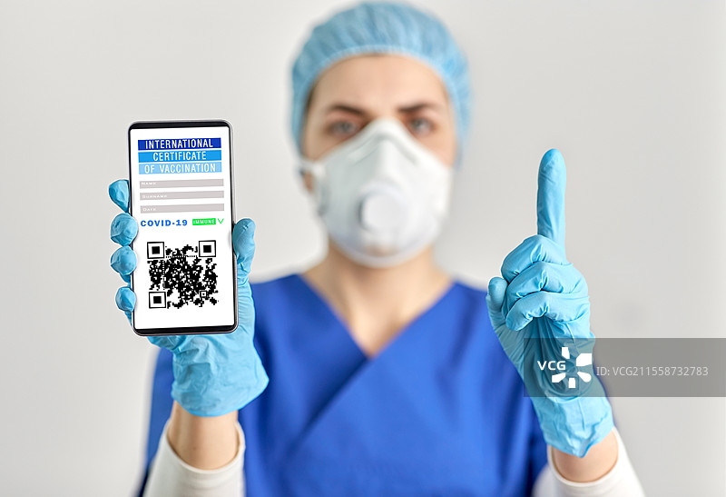 医学、健康和疫情概念 - 戴着口罩或呼吸器的医生或护士手持智能手机，上面显示虚拟免疫护照或国际疫苗接种证明，并指着上方。医生手中拿着手机中的疫苗接种证明。图片素材