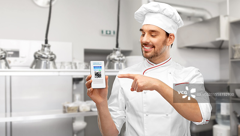 科技与健康概念 - 开心微笑的男厨师戴着厨师帽，展示着手机屏幕上的虚拟免疫护照或国际疫苗接种证明。厨师手持手机上的虚拟免疫护照。图片素材