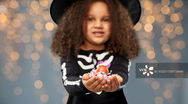 万圣节，节日和童年概念 - 非裔美国女孩穿着黑色服装和女巫帽，手里拿着糖果，在灰色背景上灯光闪烁中进行“不给糖就捣蛋”。女孩在万圣节时手里拿着糖果进行“不给糖就捣蛋”。图片素材