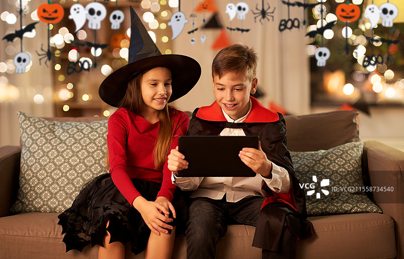 万圣节，节日和童年概念 - 微笑的男孩和女孩穿着女巫和德古拉的服装，在家中使用平板电脑。孩子们在家中穿着万圣节服装，使用平板电脑。图片素材
