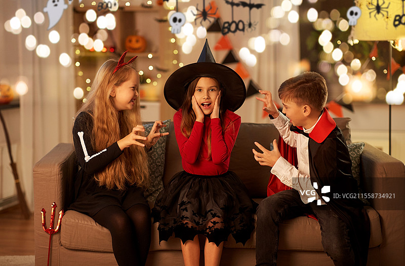 万圣节、节日和童年概念 - 笑着的男孩和女孩们穿着服装，在夜晚的家中互相捉弄，周围装饰着花环和灯光。孩子们在家中穿着万圣节服装玩耍。图片素材