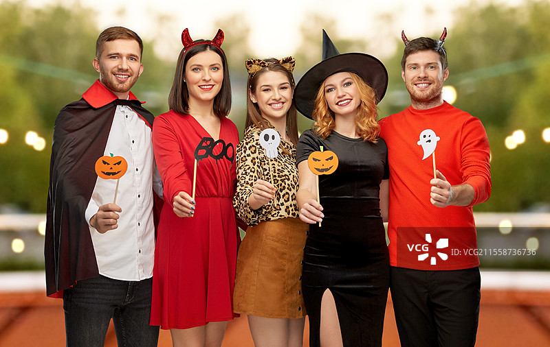 友谊、假期和照片亭概念 - 一群快乐微笑的朋友穿着恶魔、女巫和豹子的万圣节服装，手持派对道具，背景是屋顶派对。穿着万圣节服装的朋友们与派对道具。图片素材