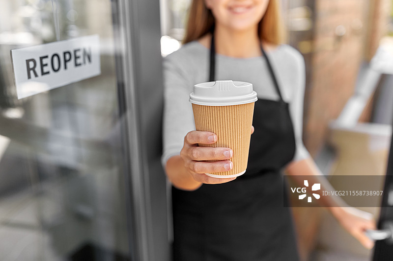 小型企业，重新开业和服务概念 - 开心微笑的女性手持外带咖啡杯，窗户或门玻璃上贴有重新开业的横幅。手握咖啡的快乐女性与门上的重新开业横幅。图片素材