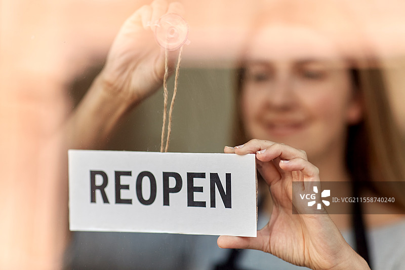 小型企业，重新开业和服务理念 - 开心微笑的女性在窗户或门玻璃上悬挂重新开业的横幅。开心的女性在门玻璃上悬挂重新开业的横幅。图片素材