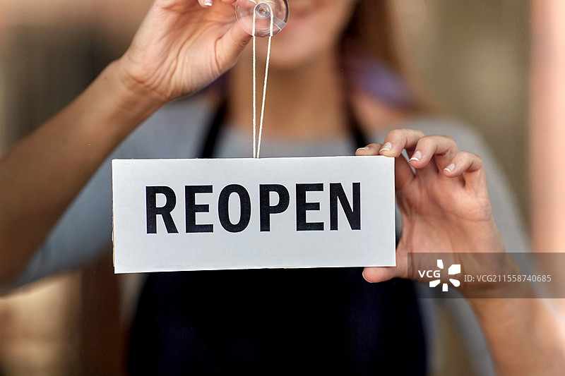 小型企业，重新开业和服务概念 - 开心微笑的女性在窗户或门玻璃上悬挂重新开业的横幅。开心的女性在门玻璃上悬挂重新开业的横幅。图片素材