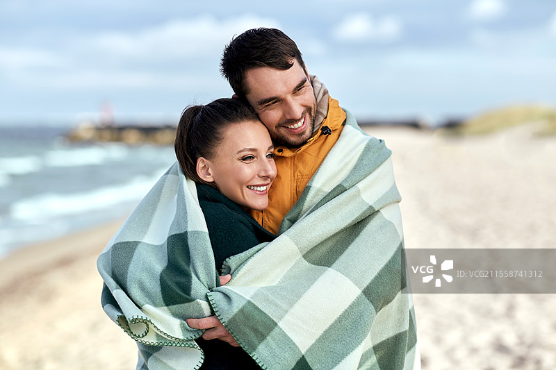爱情、关系和人际关系的概念 - 在秋季海滩上，快乐微笑的情侣裹在温暖的毛毯中。快乐的情侣在秋季海滩上裹着温暖的毛毯。图片素材