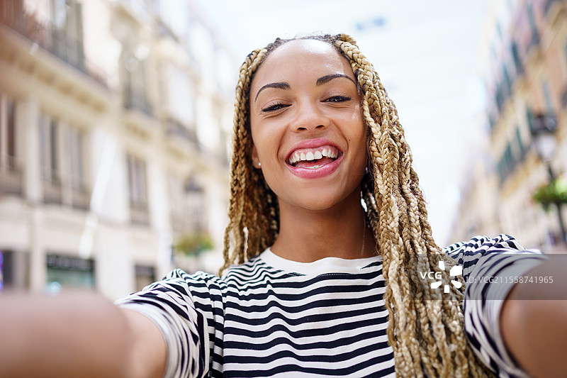 微笑的非裔美国女性，扎着非洲辫子，在城市街道上用智能手机自拍。图片素材