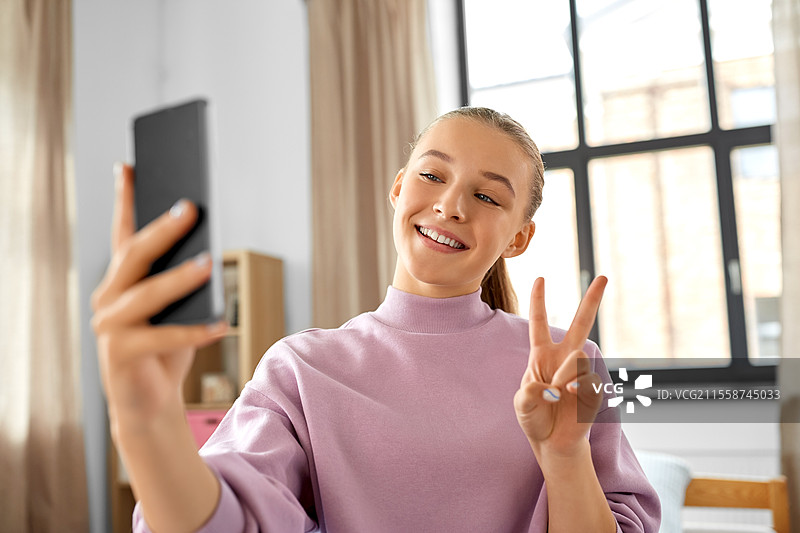 人们、儿童与科技的概念 - 开心微笑的少女在家用智能手机自拍并做出和平手势。开心的女孩在家用智能手机自拍。图片素材