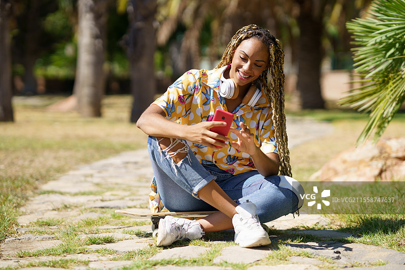 非洲女孩扎着辫子，坐在滑板上用智能手机，街道上。图片素材