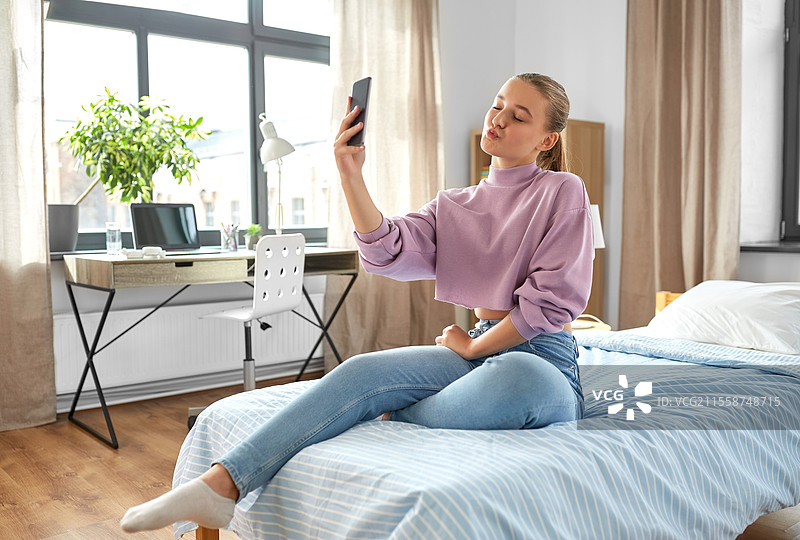 人们、儿童与科技的概念 - 开心微笑的少女在家用智能手机自拍并做鸭嘴脸。快乐的女孩在家用智能手机自拍。图片素材