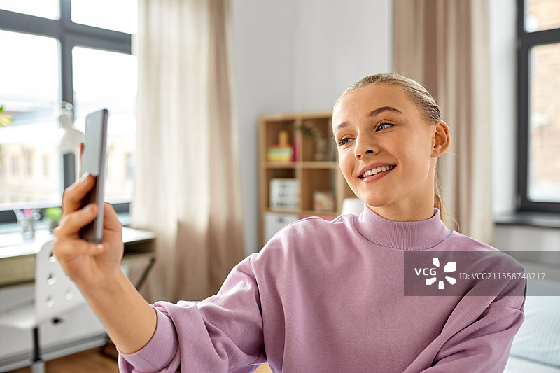 人们、儿童与科技的概念 - 开心微笑的少女在家用智能手机自拍。开心的女孩在家用智能手机自拍。图片素材