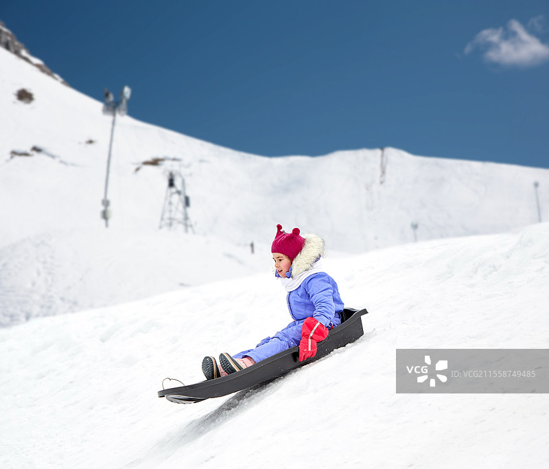 童年、滑雪和季节概念 - 快乐的小女孩在阿尔卑斯山背景下的冬季户外雪坡上滑雪橇。快乐的小女孩在冬天滑下雪橇。图片素材