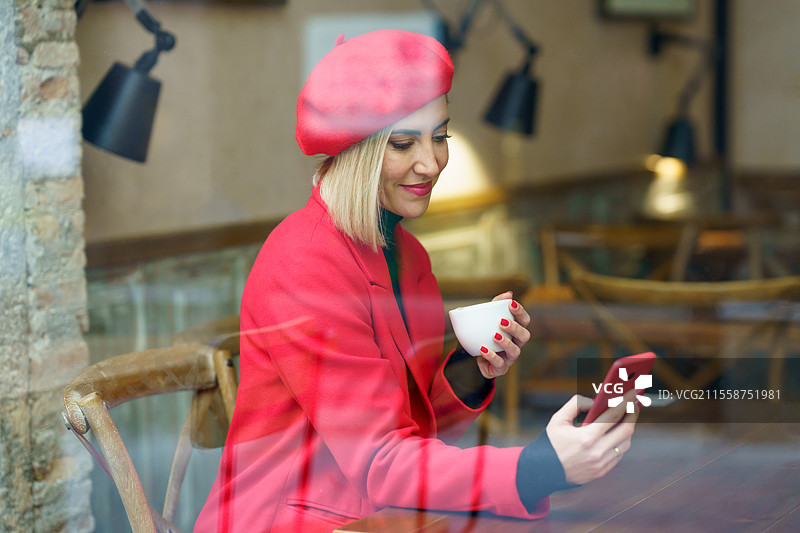 中年女性在咖啡店里喝着一杯咖啡，坐在桌子旁查看她的智能手机，穿着红色衣服透过餐厅的窗户可见。图片素材