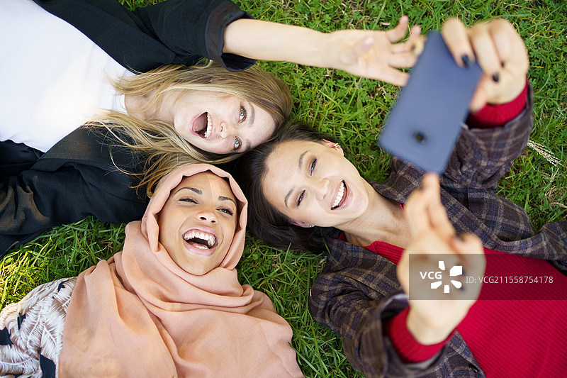 从上方看，兴奋的年轻时尚多民族女性，最好的朋友们穿着休闲服，躺在草坪上用智能手机自拍，享受在公园的时光。快乐的多元文化女孩们在公园的草坪上躺着，欢笑着自拍。图片素材