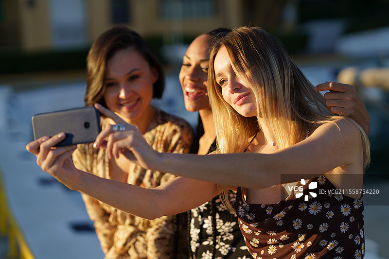 年轻的多民族女性朋友们在一起享受时光，使用手机拍摄自拍照。迷人的多样化女性朋友们在智能手机上合影。图片素材