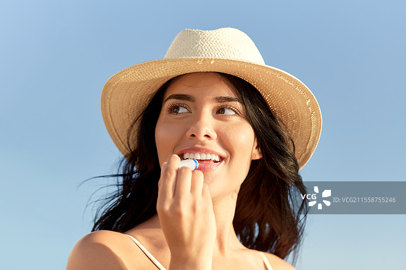 夏天和泳装概念 - 一位快乐微笑的年轻女性在海滩上穿着比基尼泳衣和草帽，涂抹润唇膏。海滩上微笑的女性与润唇膏。图片素材