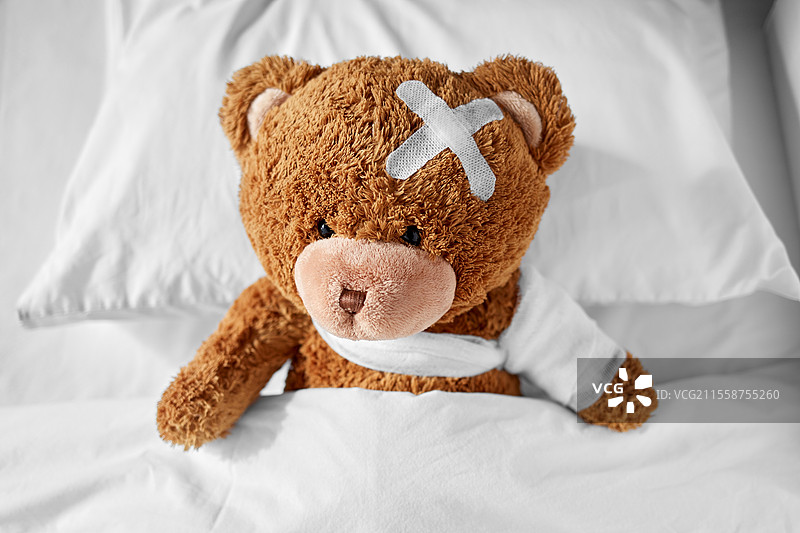 医学、医疗和儿童概念 - 生病的泰迪熊玩具，包扎着爪子，头上贴着医疗贴，躺在床上。生病的泰迪熊，爪子和头部包扎，躺在床上。图片素材