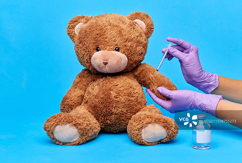 疫苗接种、医疗保健和疫情概念 - 医生戴着医疗手套的手持疫苗和注射器正在对蓝色背景上的玩具泰迪熊进行注射。玩具泰迪熊与手持疫苗和注射器的手。图片素材