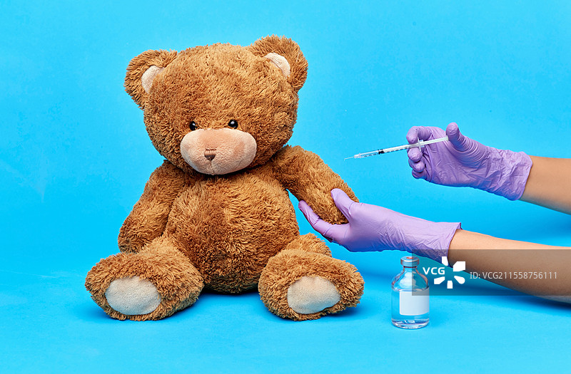 疫苗接种、医疗保健和疫情概念 - 医生戴着医疗手套的手持疫苗和注射器，正在对蓝色背景上的玩具泰迪熊进行注射。玩具泰迪熊和手持疫苗及注射器的手。图片素材