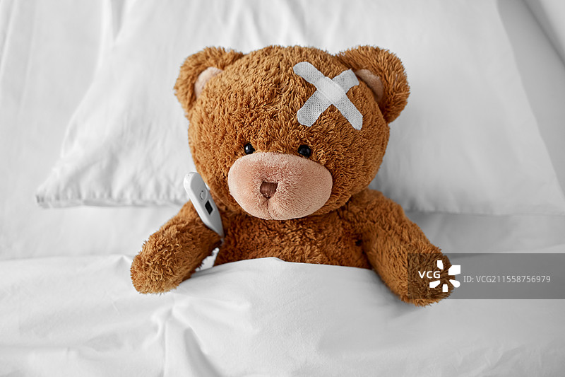 医学、医疗保健和儿童概念 - 生病的泰迪熊玩具，头上贴着医疗贴片，躺在床上，旁边有温度计。生病的泰迪熊带着贴片和温度计躺在床上。图片素材