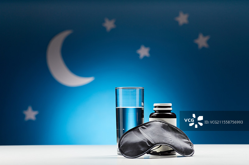 睡眠障碍，睡前和物体概念 - 近距离拍摄眼罩、水杯和安眠药，背景为蓝色的月亮和夜空星星。夜晚的水、眼罩和药物。图片素材