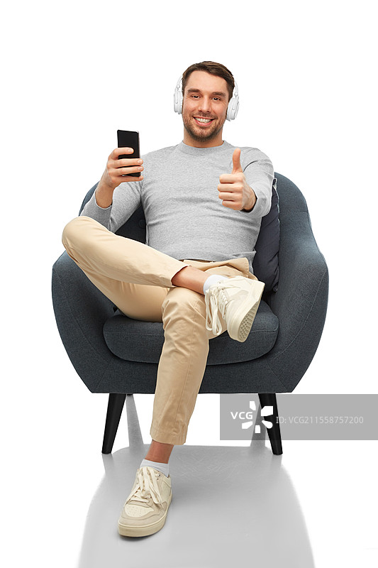 科技、人与音乐的概念 - 开心微笑的男子坐在椅子上，手持智能手机和耳机，背景为白色，竖起大拇指。男子与手机和耳机竖起大拇指。图片素材