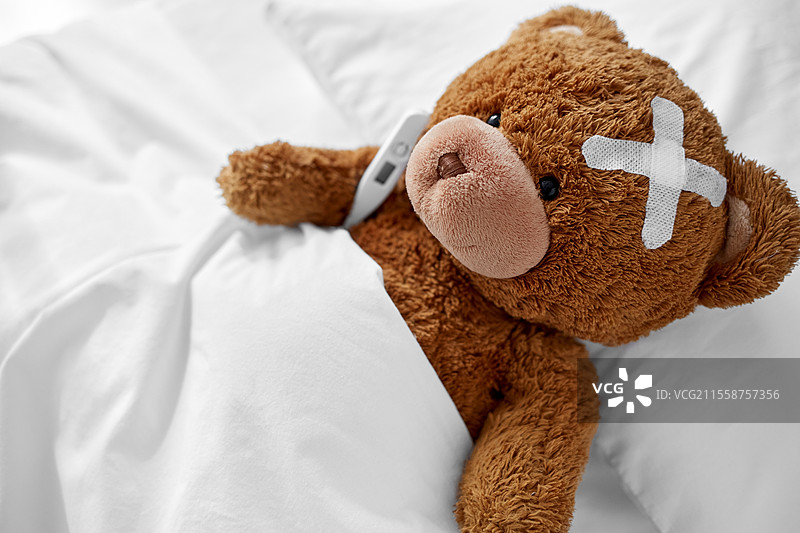医学、医疗保健与儿童概念 - 生病的泰迪熊玩具，头上贴着医疗贴片，躺在床上，旁边有温度计。生病的泰迪熊带着贴片和温度计躺在床上。图片素材