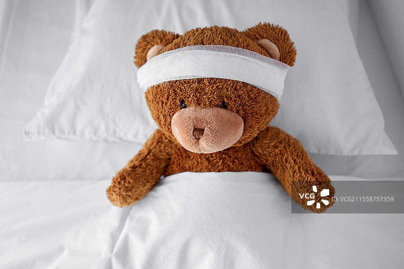 医学、医疗保健和儿童概念 - 受伤的泰迪熊玩具，头部包扎着，躺在床上。受伤的泰迪熊玩具，头部包扎着，躺在床上。图片素材