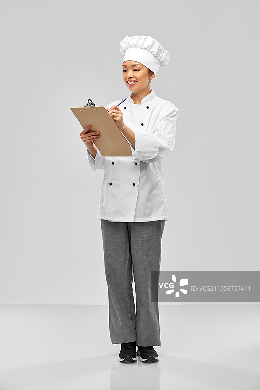 烹饪、烹饪艺术和人们的概念 - 开心微笑的女厨师手持夹板和笔，背景为灰色。微笑的女厨师手持夹板和笔。图片素材