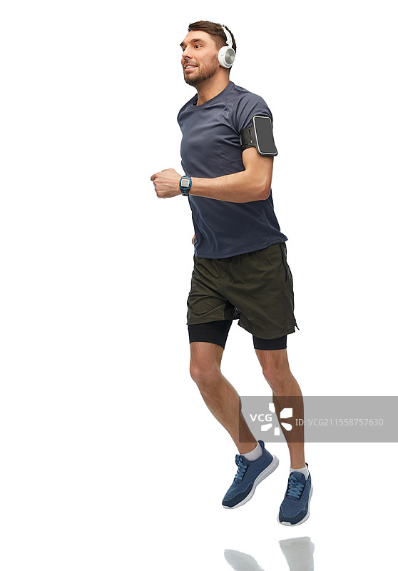 健身、运动和健康生活方式概念 - 戴着耳机、手臂上挂着智能手机的快乐微笑男子在白色背景上奔跑。戴着耳机的微笑男子在奔跑。图片素材